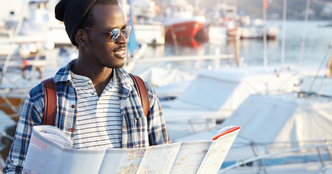 Outdoor portrait of African man looking happy before journey, waiting for his friends in harbor, holding paper map, feeling excited and joyful, anticipating adventures, places and good experience